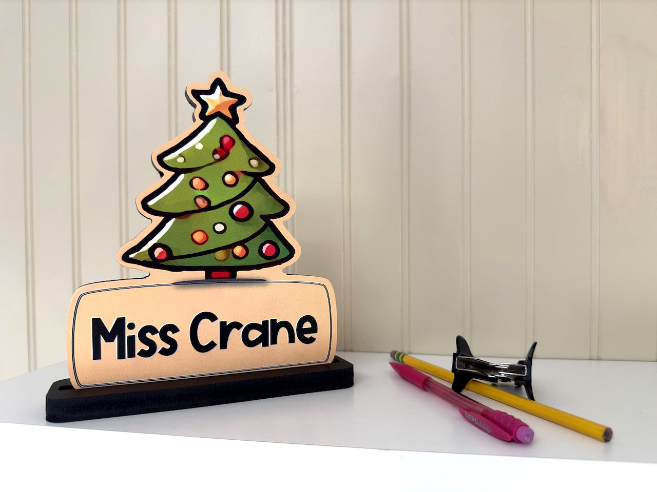 Festive Christmas tree name sign with 'Miss Crane' displayed on a desk, accompanied by office supplies like pens and a stapler.