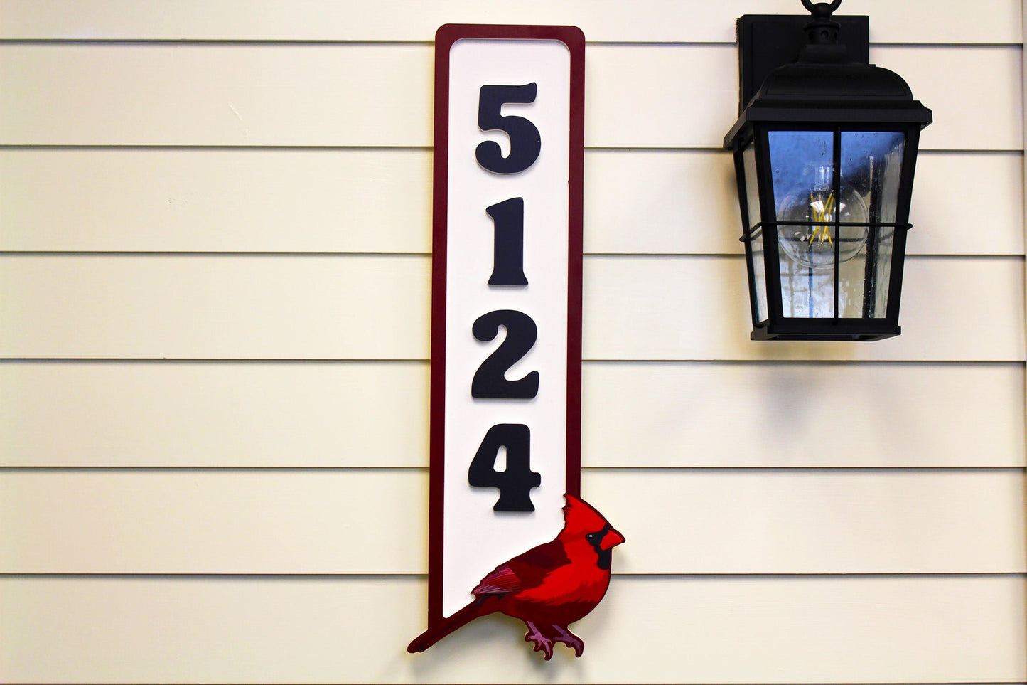 Full front view of a vertical house number sign with a red cardinal at the bottom and black numbers on a white background, mounted beside a black outdoor lamp.
