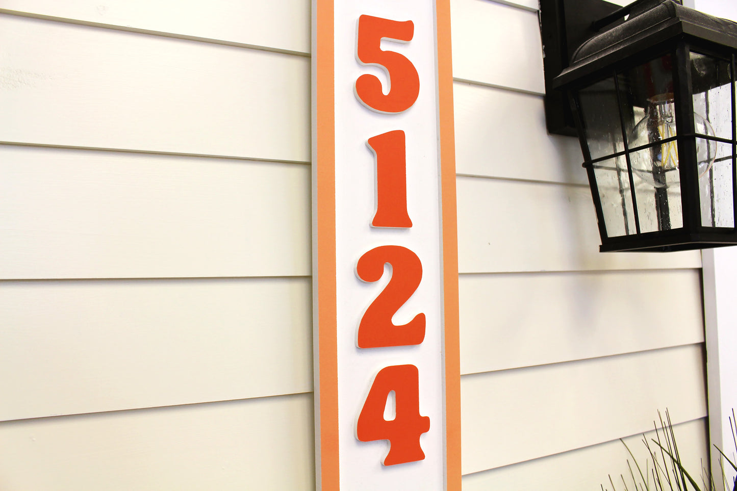 Angled view of a vertical house number sign with orange numbers and border, showing depth and detail against the wall.