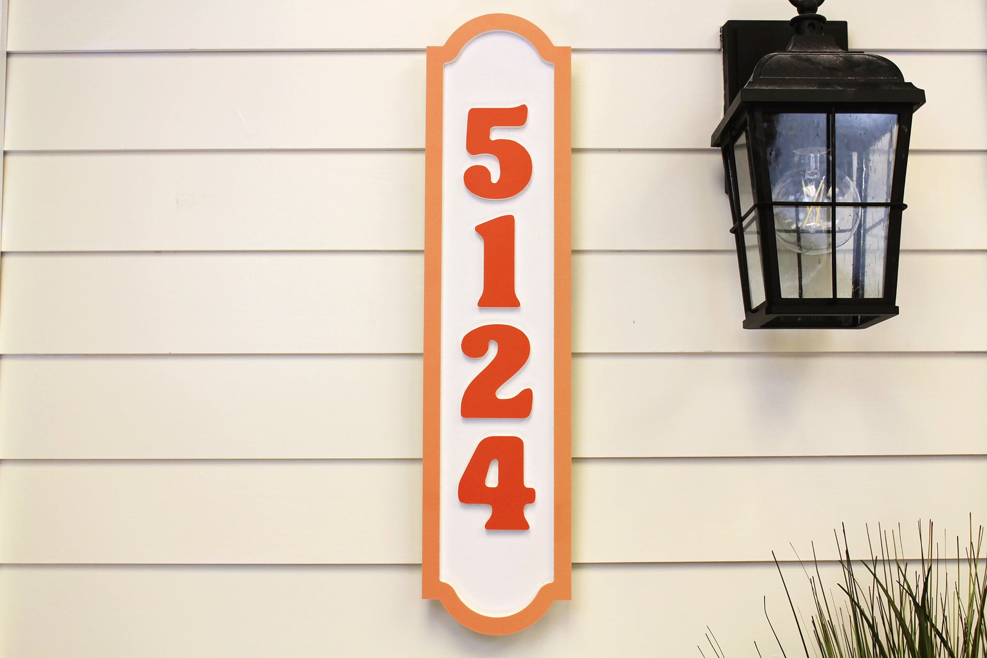 Vertical house number sign with orange numbers and border, mounted on a wall near an outdoor lamp.