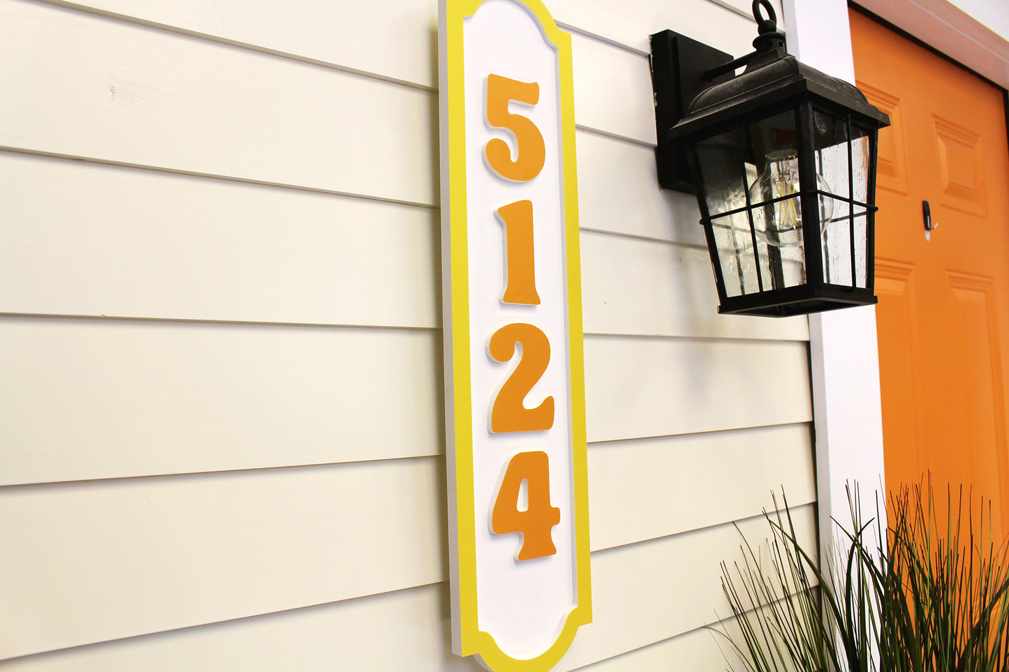 Angled side view of a vertical house number sign with yellow border and orange numbers, mounted next to a lamp on an exterior wall.