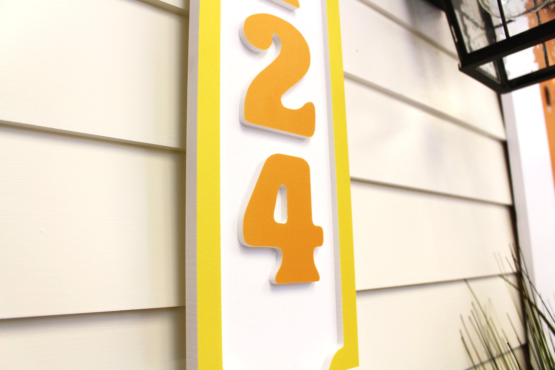 Close-up view of a yellow-bordered house number sign with raised orange numbers, showing detailed craftsmanship and texture.