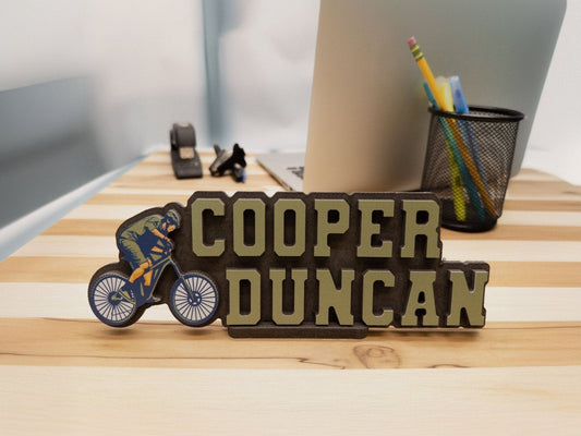 Desk name sign for Cooper Duncan featuring a mountain bike rider, displayed on an office desk.