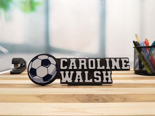 Personalized desk name sign for Caroline Walsh with a soccer ball design, displayed on a wooden desk with office supplies.