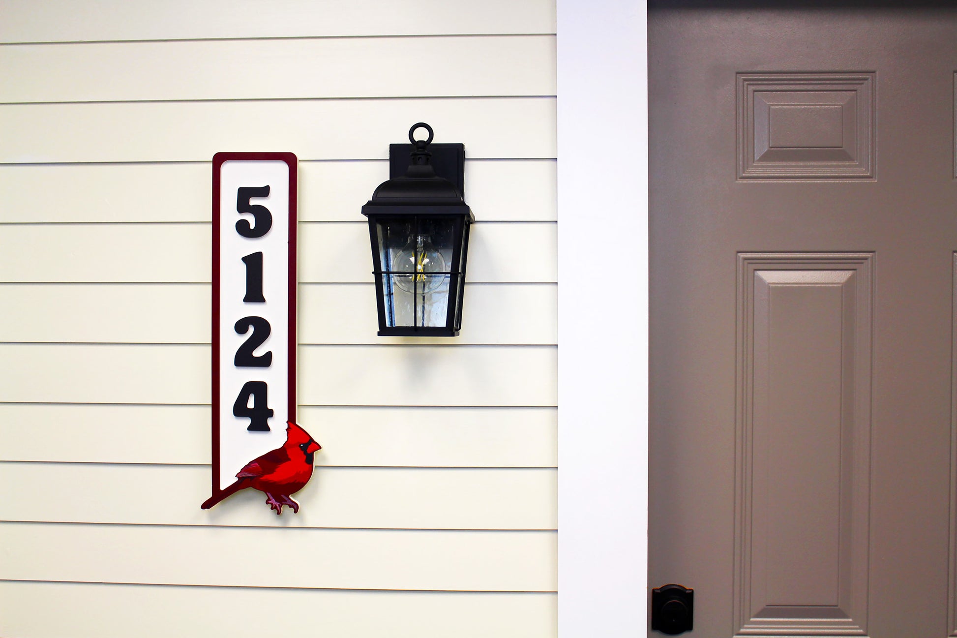 Vertical house number sign with black numbers and a red cardinal, mounted on a white and beige wall beside a black outdoor lamp